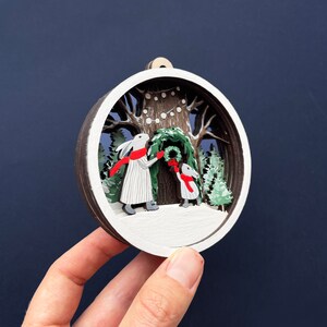May include: A white wooden ornament with a 3D scene of two white rabbits decorating a Christmas tree with red and green accents. The ornament is round and has a white background with a brown wooden frame.