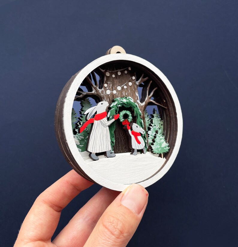 May include: A white wooden ornament with a 3D scene of two white rabbits decorating a Christmas tree with red and green accents. The ornament is round and has a white background with a brown wooden frame.