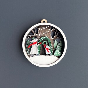 May include: A white wooden ornament with a 3D design of two rabbits hanging a wreath on a tree. The tree is decorated with white lights and the rabbits are wearing red scarves.