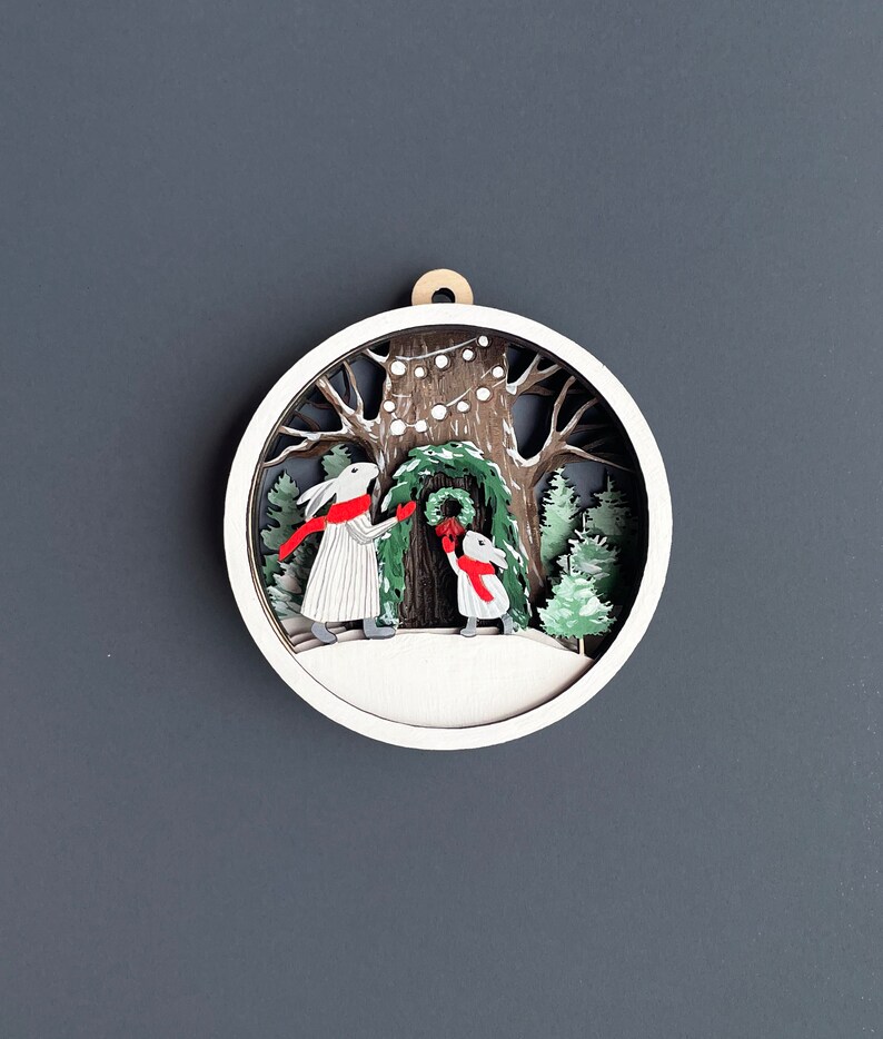 May include: A white wooden ornament with a 3D design of two rabbits hanging a wreath on a tree. The tree is decorated with white lights and the rabbits are wearing red scarves.