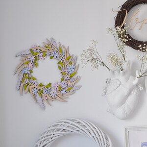 Puede incluir: Una corona de madera con flores de color rosa pastel, morado y azul y hojas verdes. La corona est&aacute; colgada en una pared blanca.