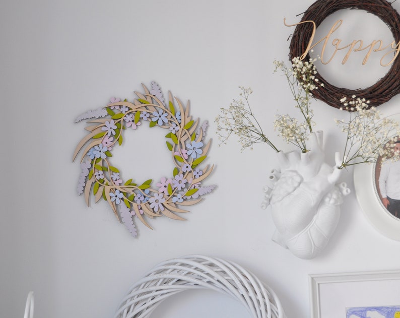 Puede incluir: Una corona de madera con flores de color rosa pastel, morado y azul y hojas verdes. La corona est&aacute; colgada en una pared blanca.
