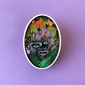 May include: A white oval frame with a paper cut-out design of three raccoons in a forest setting. The raccoons are surrounded by green ferns and brown leaves.