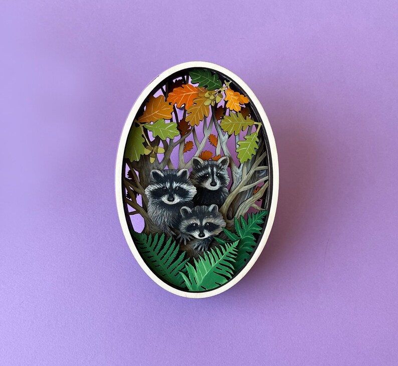 May include: A white oval frame with a paper cut-out design of three raccoons in a forest setting. The raccoons are surrounded by green ferns and brown leaves.