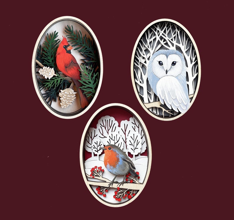 Puede incluir: Tres adornos de madera con escenas de invierno. El primer adorno representa un cardenal rojo posado en una rama con hojas verdes y pi&ntilde;as. El segundo adorno muestra un b&uacute;ho blanco con ojos negros sentado en una rama con &aacute;rboles desnudos. El tercer adorno presenta un petirrojo con el pecho rojo sentado en una rama con bayas rojas y &aacute;rboles cubiertos de nieve.