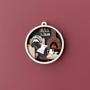 May include: A circular wooden ornament with a badger sitting in a chair by a fireplace. The badger is reading a book and there is a teapot and cup on a table in front of the fireplace.