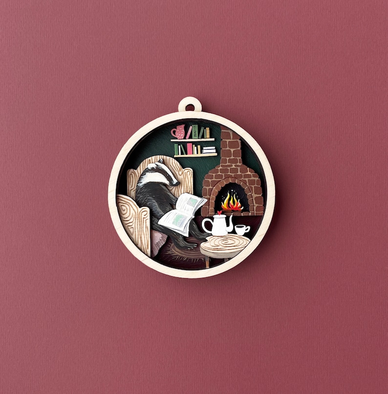 May include: A circular wooden ornament with a badger sitting in a chair by a fireplace. The badger is reading a book and there is a teapot and cup on a table in front of the fireplace.