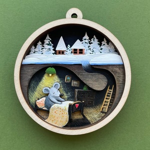 Może przedstawiać: Drewniana ozdoba przedstawiająca mysz w przytulnym łóżku z poduszką w kształcie czerwonego serca, osadzoną w śnieżnym zimowym krajobrazie z dwoma domami i krętym szlakiem prowadzącym do drabiny.