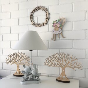 May include: Two wooden tree sculptures with intricate branches, one with a pair of white ceramic birds perched on a branch. A white lamp with a white lampshade sits on a white table, along with a wooden wreath and a wooden chair with a floral bouquet.