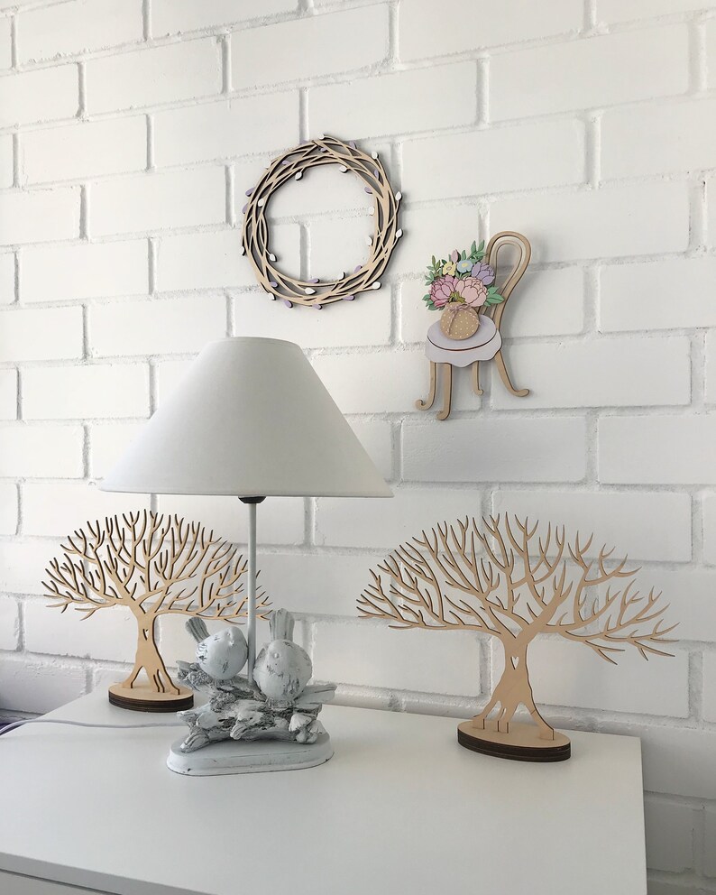 May include: Two wooden tree sculptures with intricate branches, one with a pair of white ceramic birds perched on a branch. A white lamp with a white lampshade sits on a white table, along with a wooden wreath and a wooden chair with a floral bouquet.