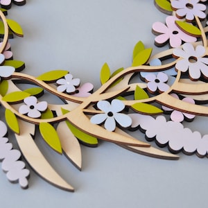 Puede incluir: Una corona de madera decorada con flores superpuestas en tonos rosa, azul y blanco. La corona est&aacute; hecha de madera natural y tiene un aspecto r&uacute;stico y hecho a mano.