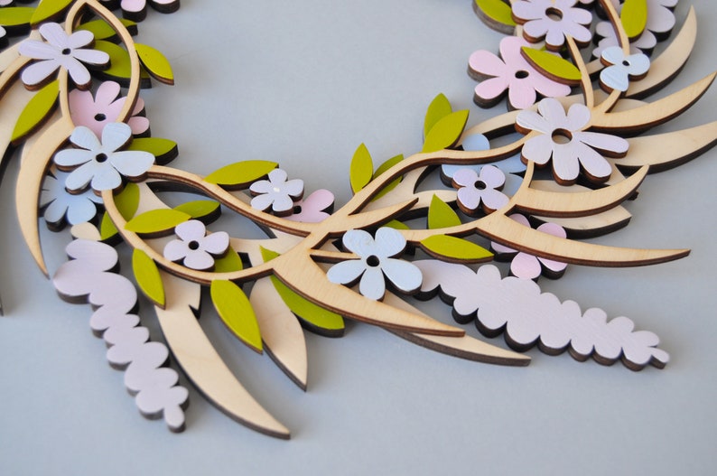 Puede incluir: Una corona de madera decorada con flores superpuestas en tonos rosa, azul y blanco. La corona est&aacute; hecha de madera natural y tiene un aspecto r&uacute;stico y hecho a mano.