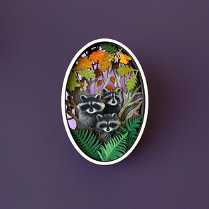May include: A paper cut-out art piece depicting three raccoons in a forest setting. The raccoons are surrounded by green ferns and brown leaves. The art piece is framed in a white oval.