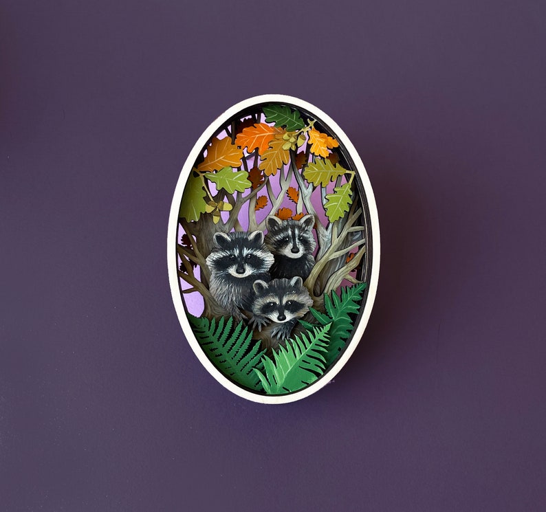 May include: A paper cut-out art piece depicting three raccoons in a forest setting. The raccoons are surrounded by green ferns and brown leaves. The art piece is framed in a white oval.