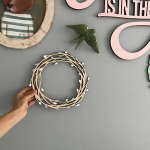 May include: A wooden wreath with a light brown base and purple and white accents. The wreath is hanging on a gray wall.