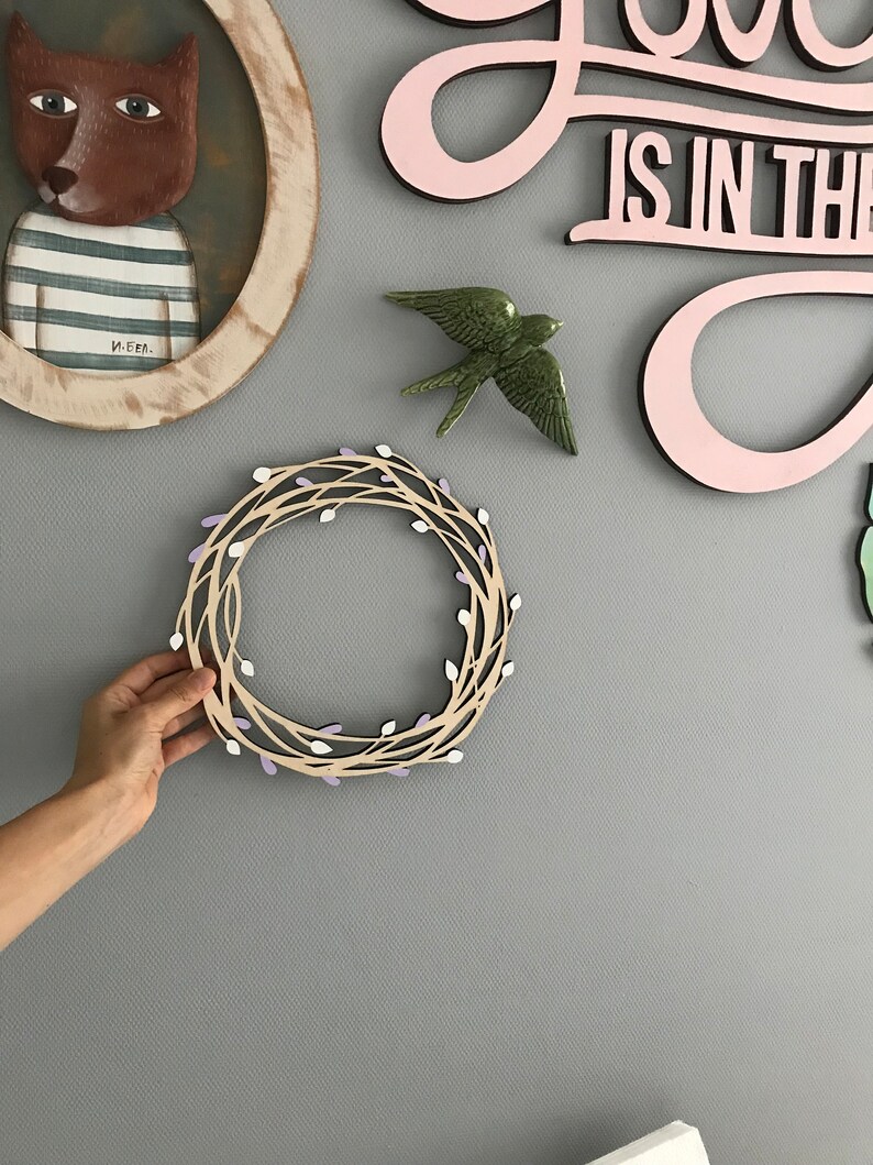 May include: A wooden wreath with a light brown base and purple and white accents. The wreath is hanging on a gray wall.