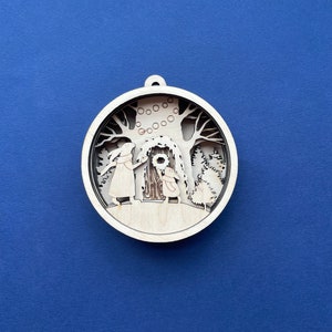 May include: A wooden Christmas ornament featuring a detailed silhouette of two figures decorating a tree with a wreath. The ornament is circular and has a rustic, hand-crafted look.