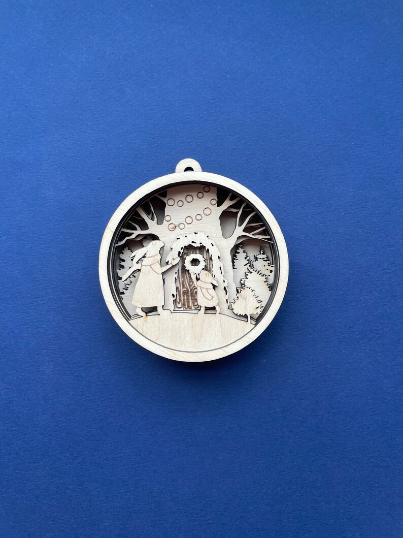 May include: A wooden Christmas ornament featuring a detailed silhouette of two figures decorating a tree with a wreath. The ornament is circular and has a rustic, hand-crafted look.