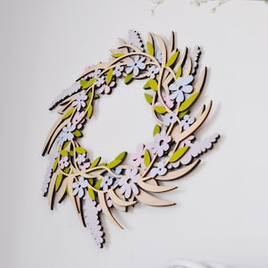 Puede incluir: Una corona de madera decorada con flores de color rosa pastel, azul y verde. La corona est&aacute; hecha de madera fina cortada con l&aacute;ser y tiene un acabado de madera natural.