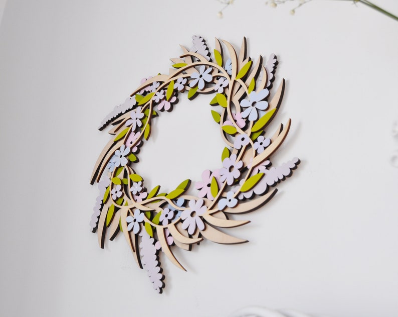 Puede incluir: Una corona de madera decorada con flores de color rosa pastel, azul y verde. La corona est&aacute; hecha de madera fina cortada con l&aacute;ser y tiene un acabado de madera natural.