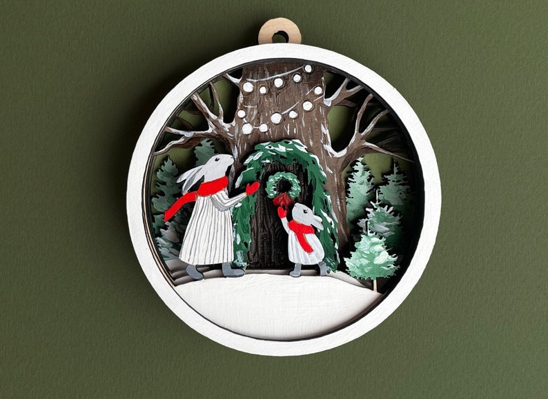 May include: A white wooden ornament with a 3D paper cut-out design of two rabbits decorating a tree with a wreath. The rabbits are wearing red scarves and the tree is decorated with white lights.