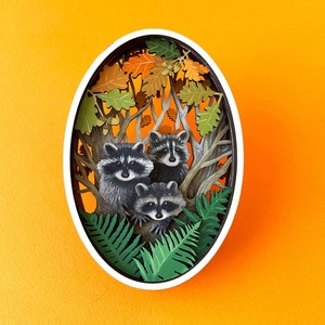 May include: A paper cut-out of three raccoons in a forest setting. The raccoons are surrounded by green leaves and brown branches. The background is a light orange color.