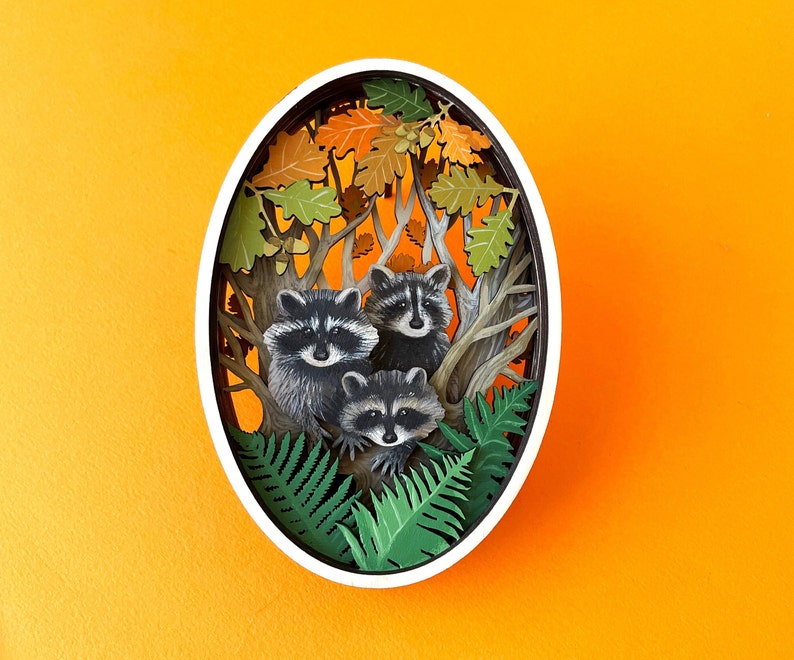 May include: A paper cut-out of three raccoons in a forest setting. The raccoons are surrounded by green leaves and brown branches. The background is a light orange color.