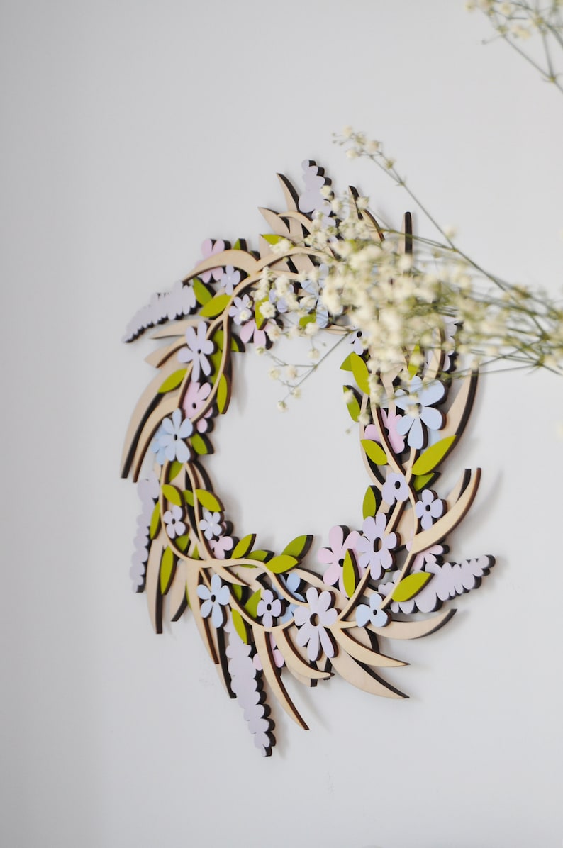 Puede incluir: Una corona de madera con un dise&ntilde;o floral. La corona est&aacute; hecha de madera marr&oacute;n claro y presenta delicadas flores en tonos de rosa, azul y verde.