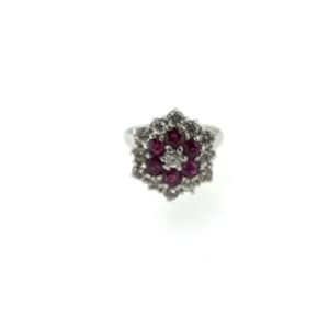 May include: A silver ring featuring a cluster of red gemstones surrounded by smaller clear stones. The ring's central design is a flower-like arrangement, with a single clear stone at the center. The ring is set against a white background.