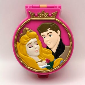 May include: A pink, clamshell-shaped toy featuring a gold-trimmed oval with a sleeping princess and a prince. The toy has a gold crown at the top and a red button at the bottom. The princess has long yellow hair and the prince has brown hair.