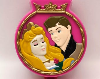 Può includere: Un giocattolo rosa a forma di conchiglia con un ovale bordato d'oro raffigurante una principessa addormentata e un principe. Il giocattolo ha una corona d'oro in alto e un bottone rosso in basso. La principessa ha lunghi capelli biondi e il principe ha i capelli castani.