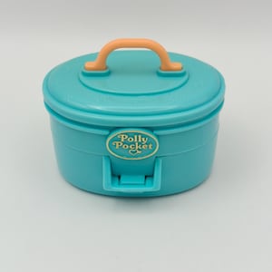 Op de afbeelding: Een turquoise Polly Pocket draagtas met een oranje handvat. De behuizing is rond met een deksel en een grendel. Het Polly Pocket-logo staat op de voorkant van de behuizing.