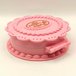 May include: A pink, round Polly Pocket compact toy. The compact is shaped like a cake with a scalloped edge and a gold-colored oval with the words "Polly Pocket" on the top. The compact has a hinged closure.