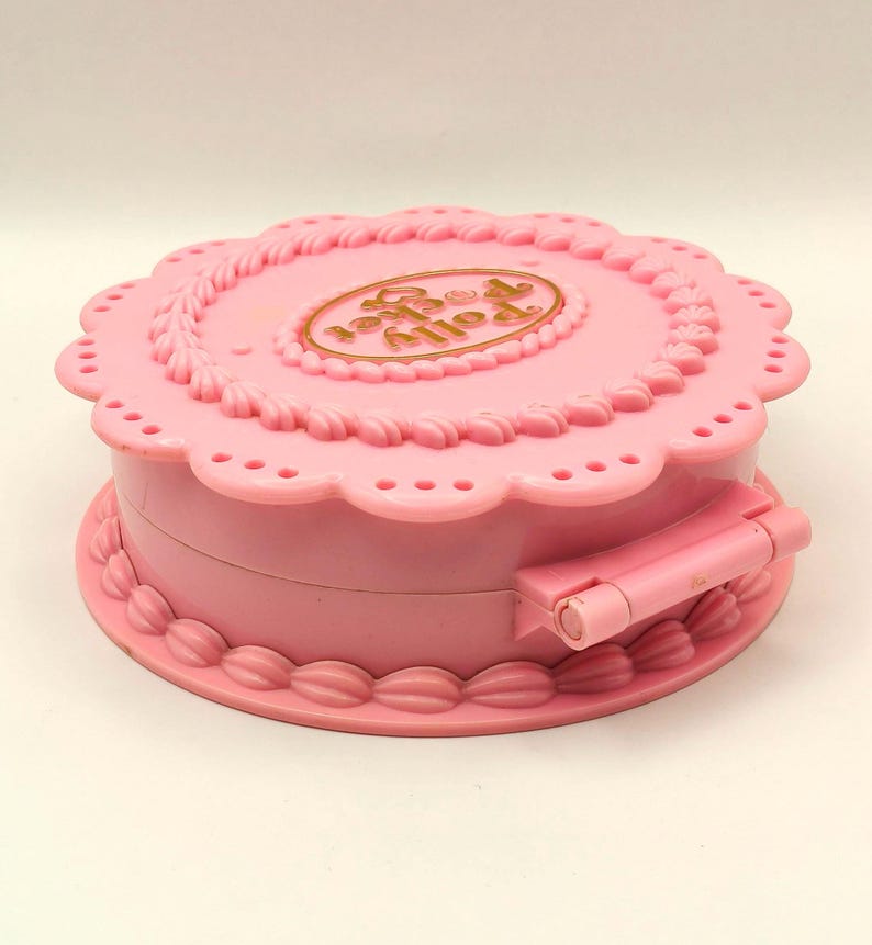 May include: A pink, round Polly Pocket compact toy. The compact is shaped like a cake with a scalloped edge and a gold-colored oval with the words "Polly Pocket" on the top. The compact has a hinged closure.