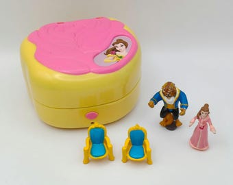 May include: A yellow and pink toy playset featuring a Beauty and the Beast theme. Includes a yellow box with a pink rose design, miniature figures of Belle, the Beast, and two chairs. A pink button is on the front.