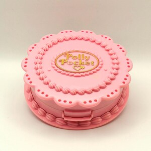 May include: A pink Polly Pocket compact in the shape of a cake. The compact has a scalloped edge and decorative frosting details. The words "Polly Pocket" are in gold lettering on the top.