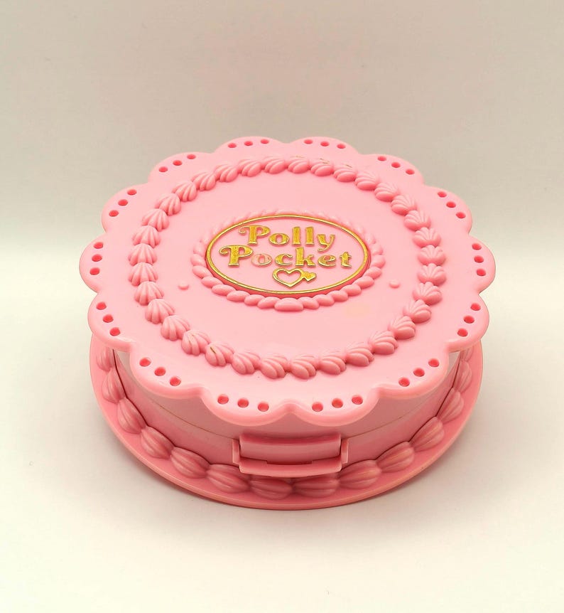 May include: A pink Polly Pocket compact in the shape of a cake. The compact has a scalloped edge and decorative frosting details. The words "Polly Pocket" are in gold lettering on the top.