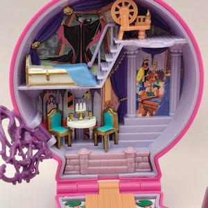 May include: A vintage pink and purple toy playset with interior scenes. The playset includes miniature furniture, stairs, and character illustrations. The top level has a spinning wheel. The interior has a bed, table, chairs, and a mural of a villain.