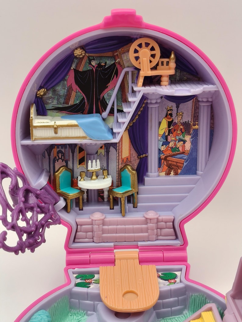 May include: A vintage pink and purple toy playset with interior scenes. The playset includes miniature furniture, stairs, and character illustrations. The top level has a spinning wheel. The interior has a bed, table, chairs, and a mural of a villain.