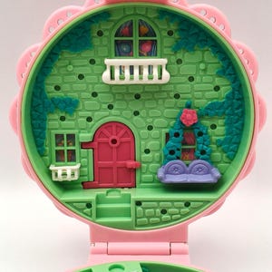 May include: A vintage pink and green miniature dollhouse with a floral design. The dollhouse features a pink door, a balcony, and a purple couch. The exterior is green with a brick pattern.