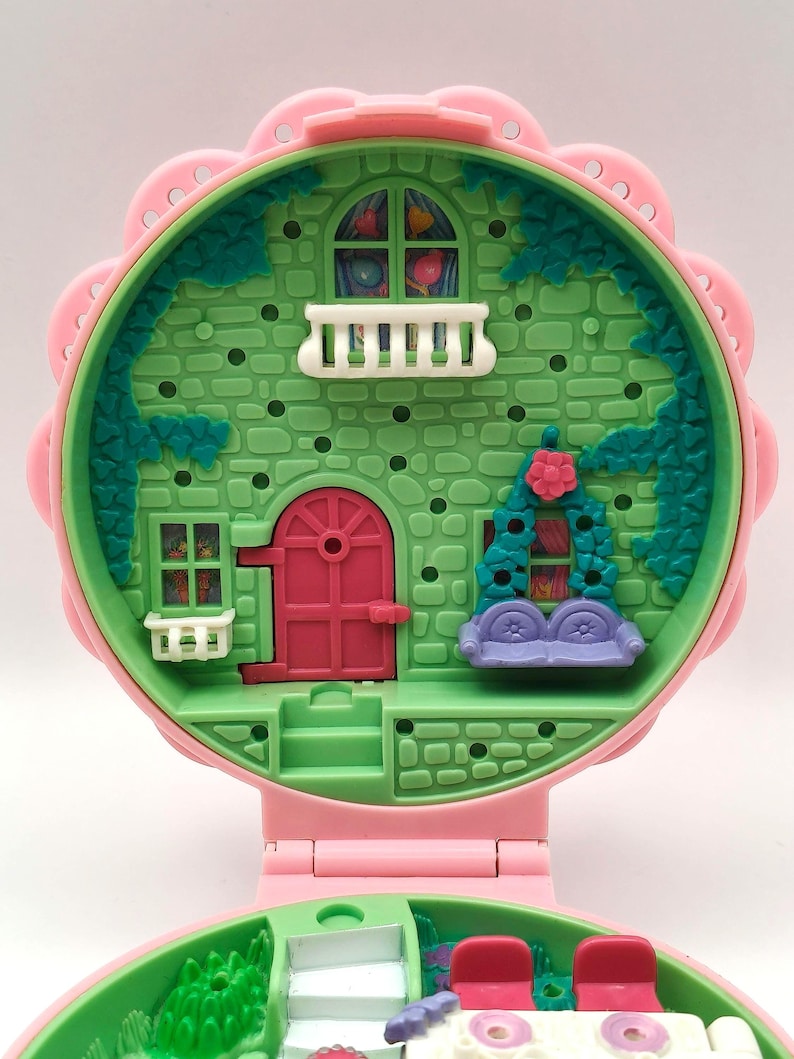 May include: A vintage pink and green miniature dollhouse with a floral design. The dollhouse features a pink door, a balcony, and a purple couch. The exterior is green with a brick pattern.