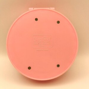 May include: A pink, circular compact with the Bluebird logo and text. The compact is closed, showing the back with four screws. The text "MADE IN CHINA" is visible. The compact is a vintage toy.