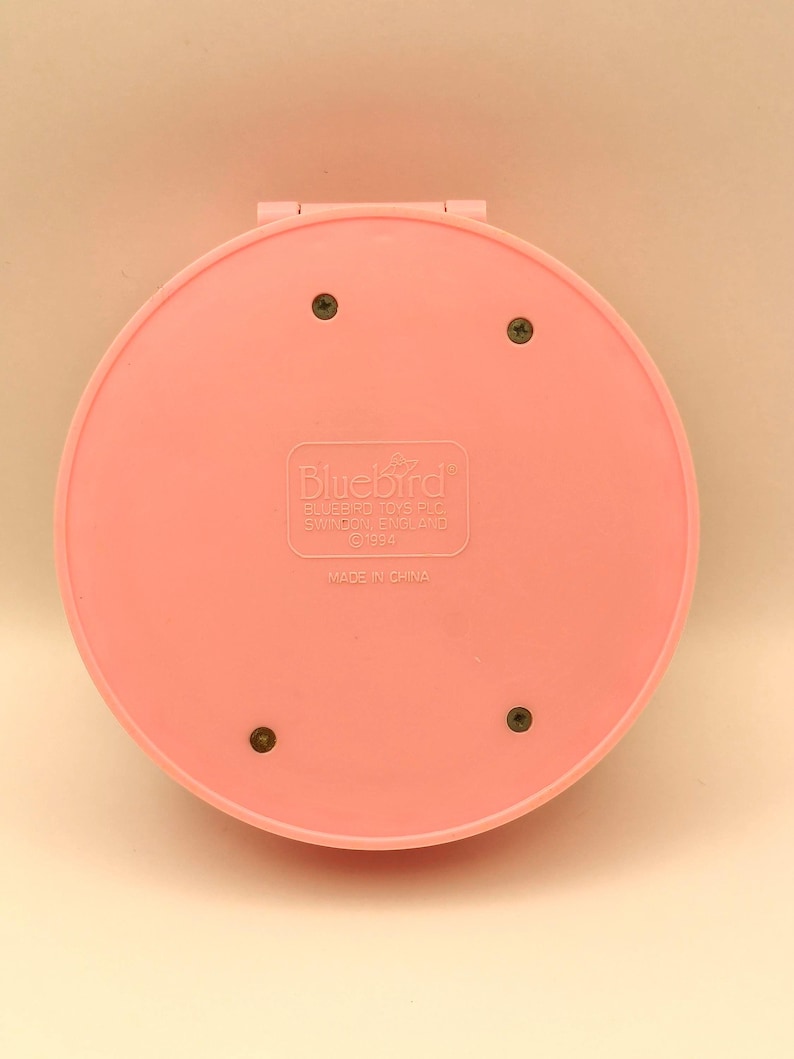 May include: A pink, circular compact with the Bluebird logo and text. The compact is closed, showing the back with four screws. The text "MADE IN CHINA" is visible. The compact is a vintage toy.