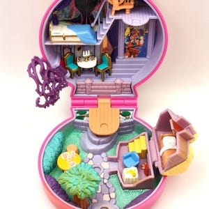 May include: A vintage pink and purple miniature playset, designed as a castle with various rooms and accessories. The playset includes a bed, table, chairs, and a garden area. The interior has a painted backdrop of a castle scene.