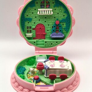 May include: A vintage pink and green miniature dollhouse, open to reveal two levels. The top level features a green facade with a red door and white balcony. The bottom level has a table with chairs and a small garden with a cat.