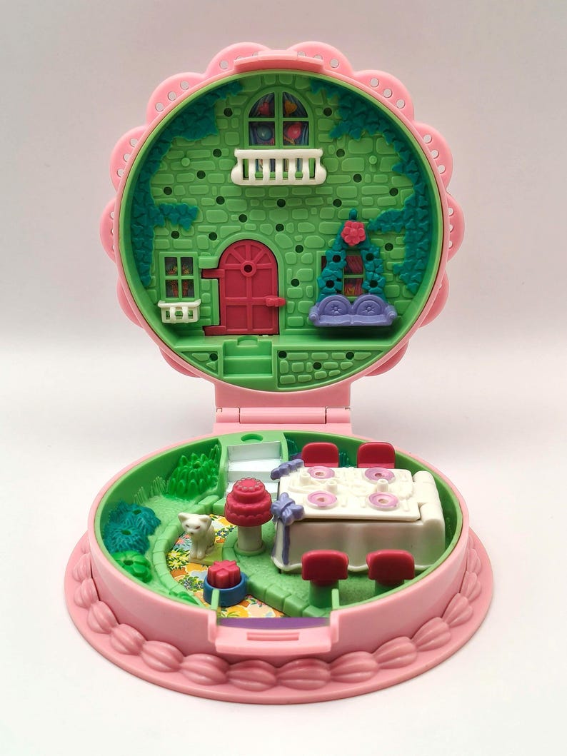 May include: A vintage pink and green miniature dollhouse, open to reveal two levels. The top level features a green facade with a red door and white balcony. The bottom level has a table with chairs and a small garden with a cat.