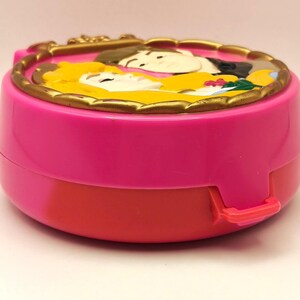 May include: A vintage pink and red compact with a gold-coloured, ornate border. The top features a relief of two figures with yellow and black hair. The compact has a clasp on the side.