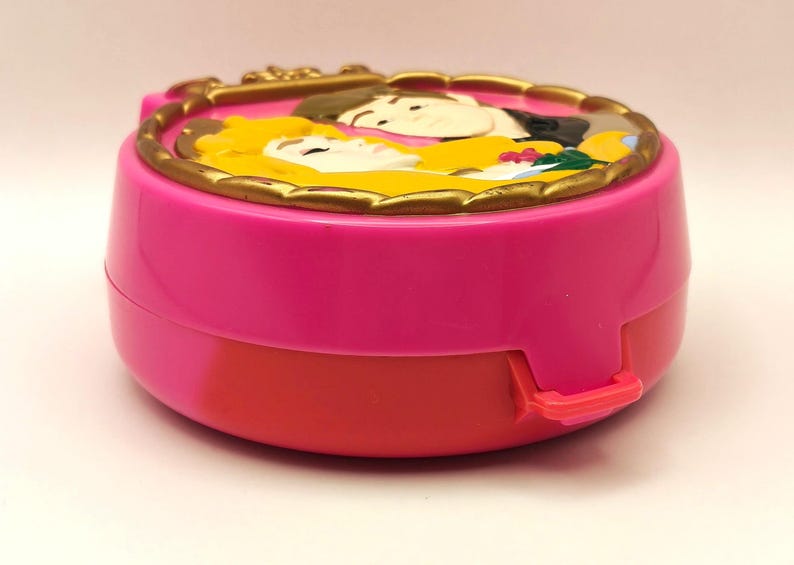 May include: A vintage pink and red compact with a gold-coloured, ornate border. The top features a relief of two figures with yellow and black hair. The compact has a clasp on the side.