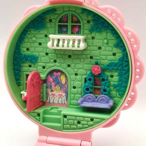 May include: A vintage, pink and green miniature dollhouse with a flower-shaped exterior. The dollhouse features a pink door, a white balcony, and a purple couch. The interior is decorated with a table and a window with a colorful design.