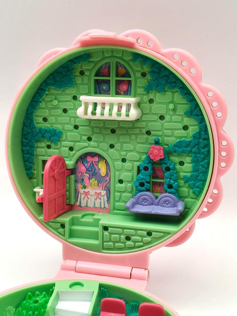 May include: A vintage, pink and green miniature dollhouse with a flower-shaped exterior. The dollhouse features a pink door, a white balcony, and a purple couch. The interior is decorated with a table and a window with a colorful design.